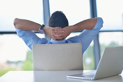 Mann lehnt sich entspannt mit den Händen hinter dem Kopf vor einem Laptop am Fenster zurück